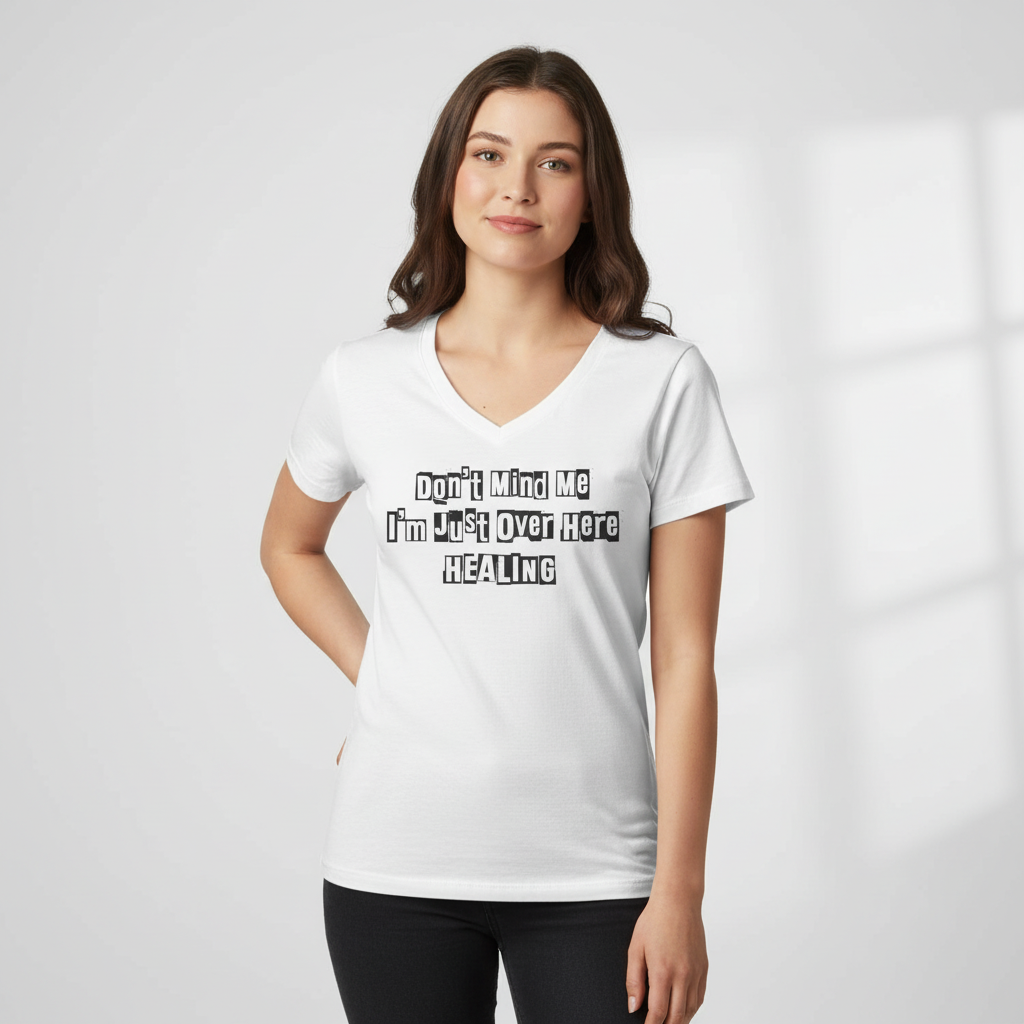 Young woman wearing a white JohnT V-neck T-shirt with the printed text “Don’t Mind Me I’m Just Over Here Healing,” standing in a minimal studio setting.