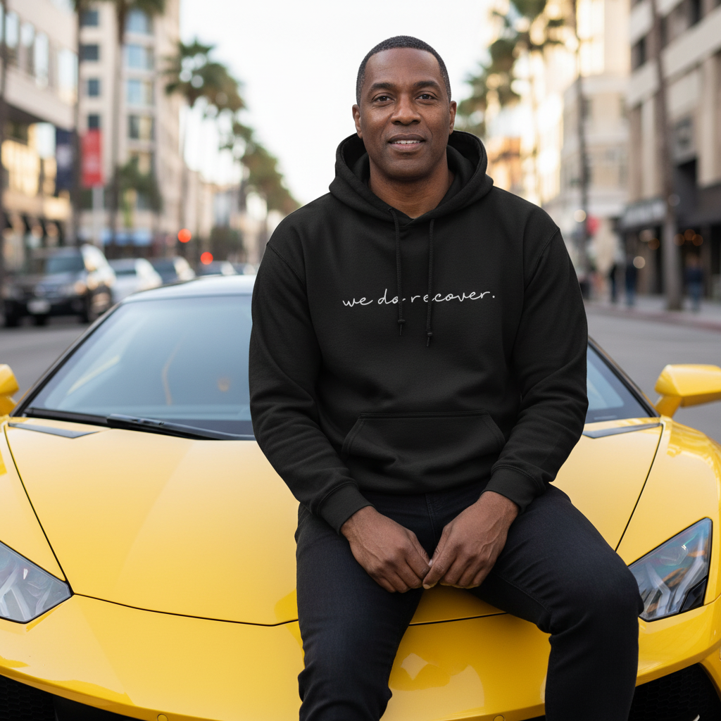 Man sitting on a yellow sports car wearing a black hoodie with white text that reads “we do recover.” representing confidence, strength, and the power of recovery