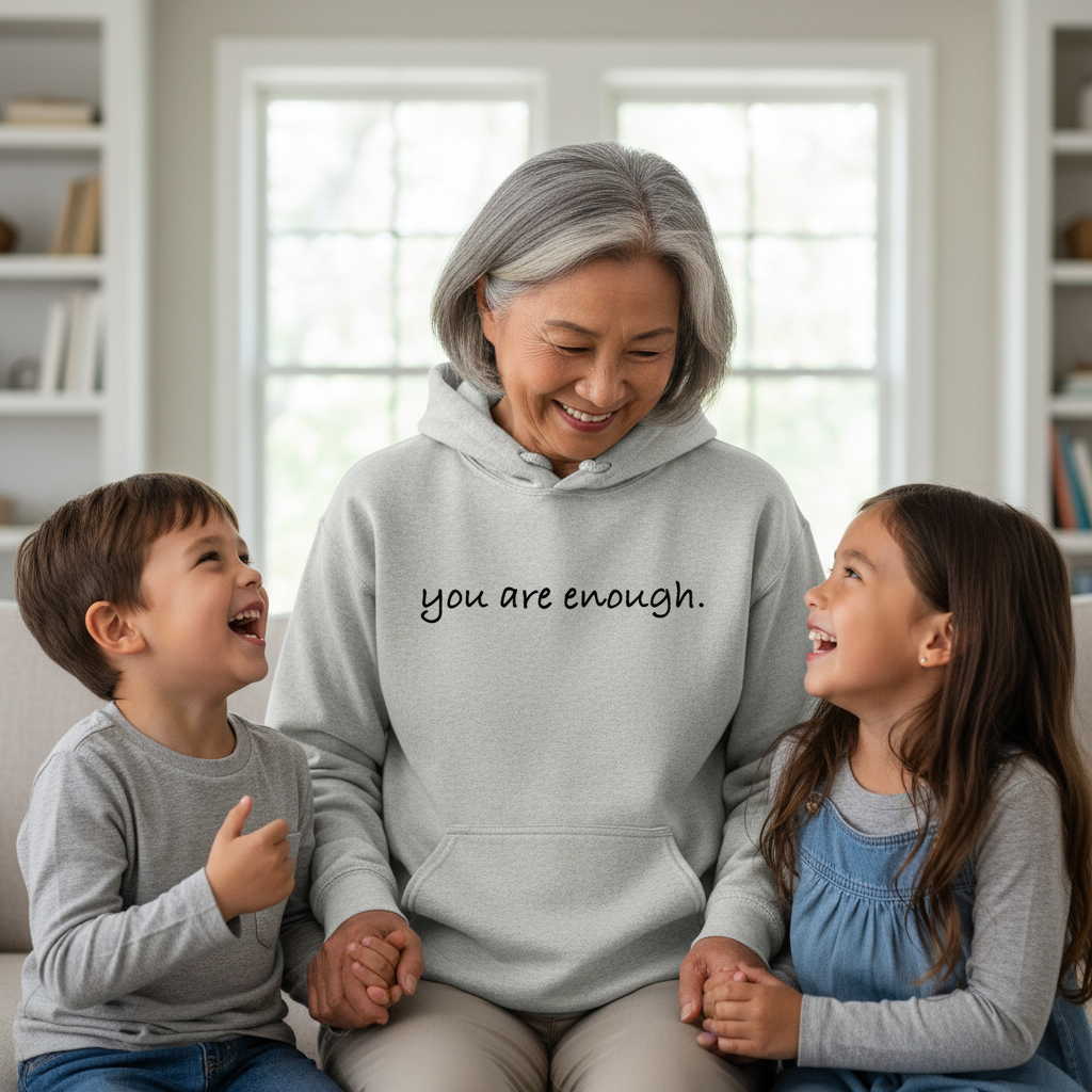 Smiling grandmother sitting with her two grandchildren, wearing a cozy grey hoodie with the text "you are enough." A warm and emotional family scene symbolizing love, reassurance, and belonging.